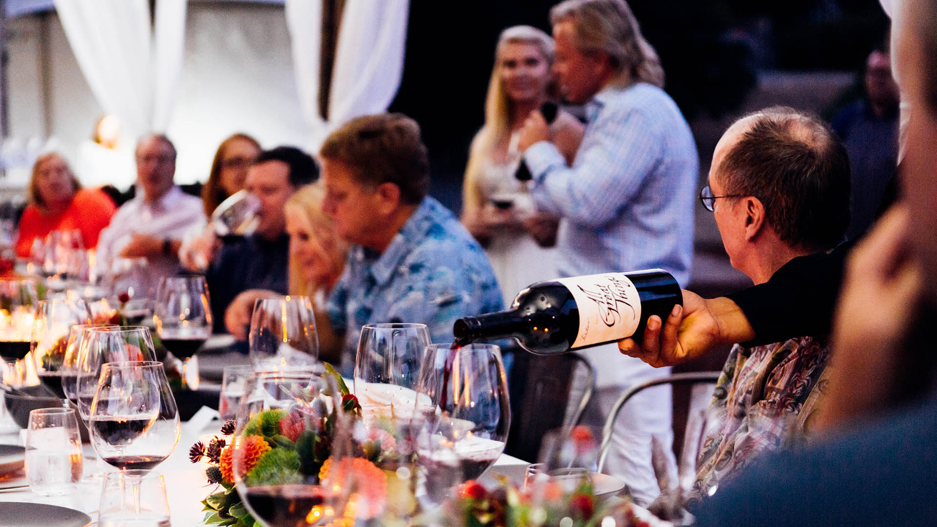 Large group of people at outdoor dining table with food and wine.